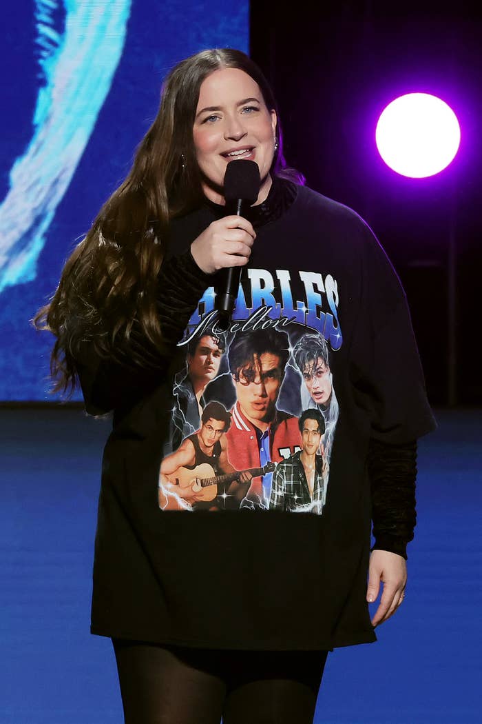 Aidy standing on stage with a microphone, wearing a sweater with a graphic print showing off Charles Melton