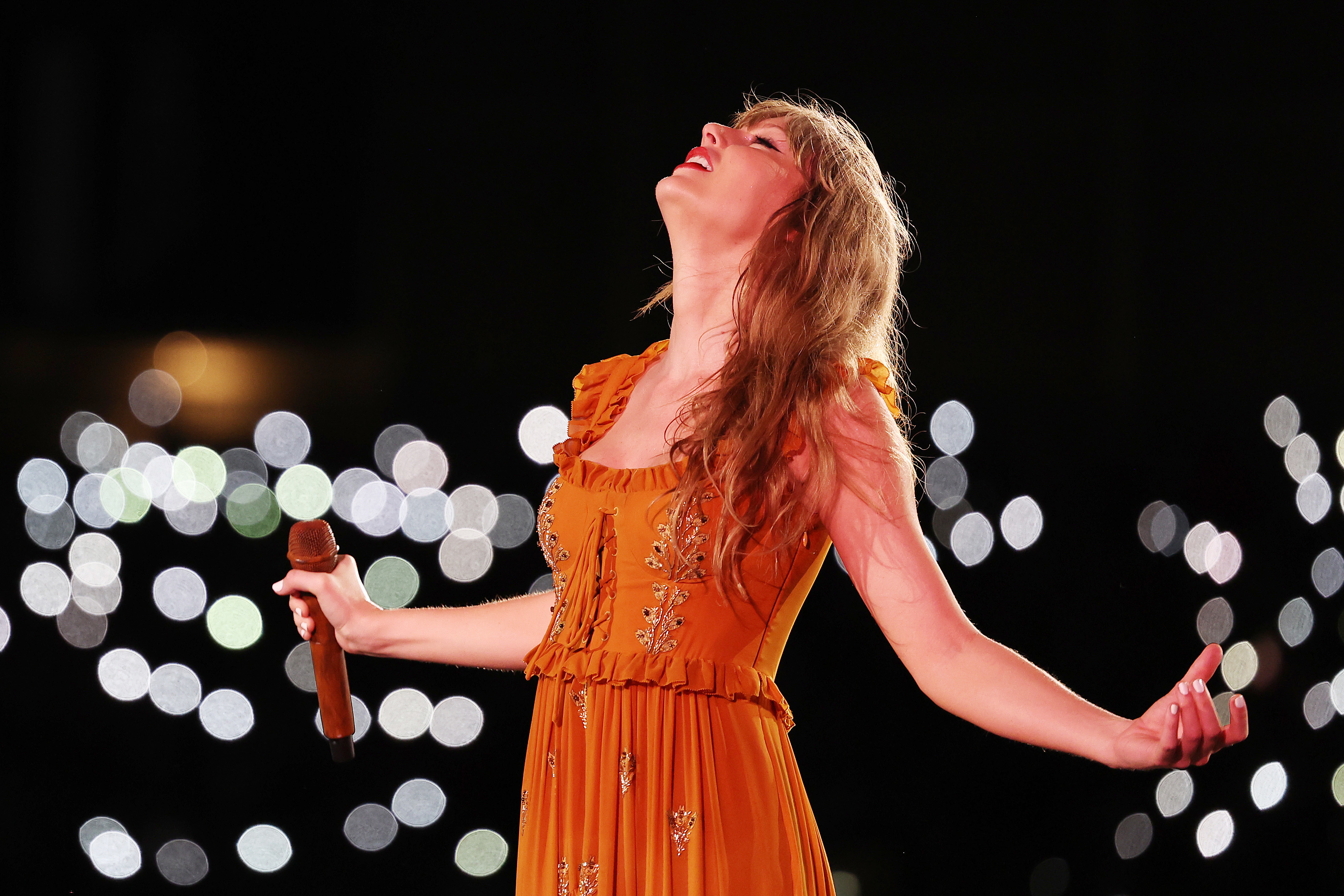Person on stage with microphone in a ruffled dress, arms outstretched in a performance pose