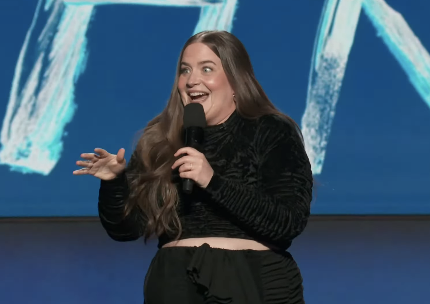 Aidy Bryant on stage at the 2022 Film Independent Spirit Awards, speaking into a microphone with projected graphics behind her