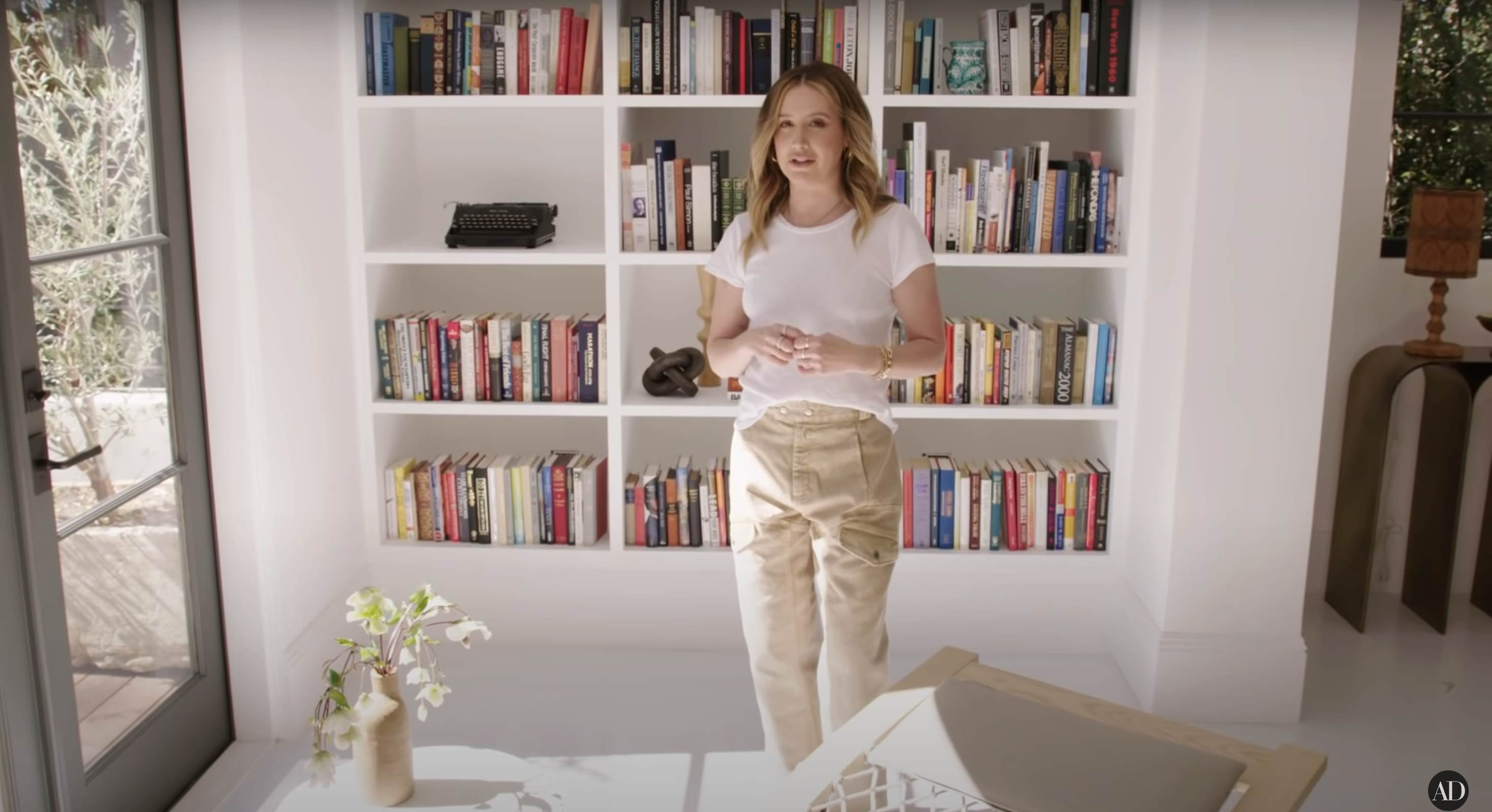 Ashley standing by a desk with a bookshelf behind her, wearing a casual top and satin trousers