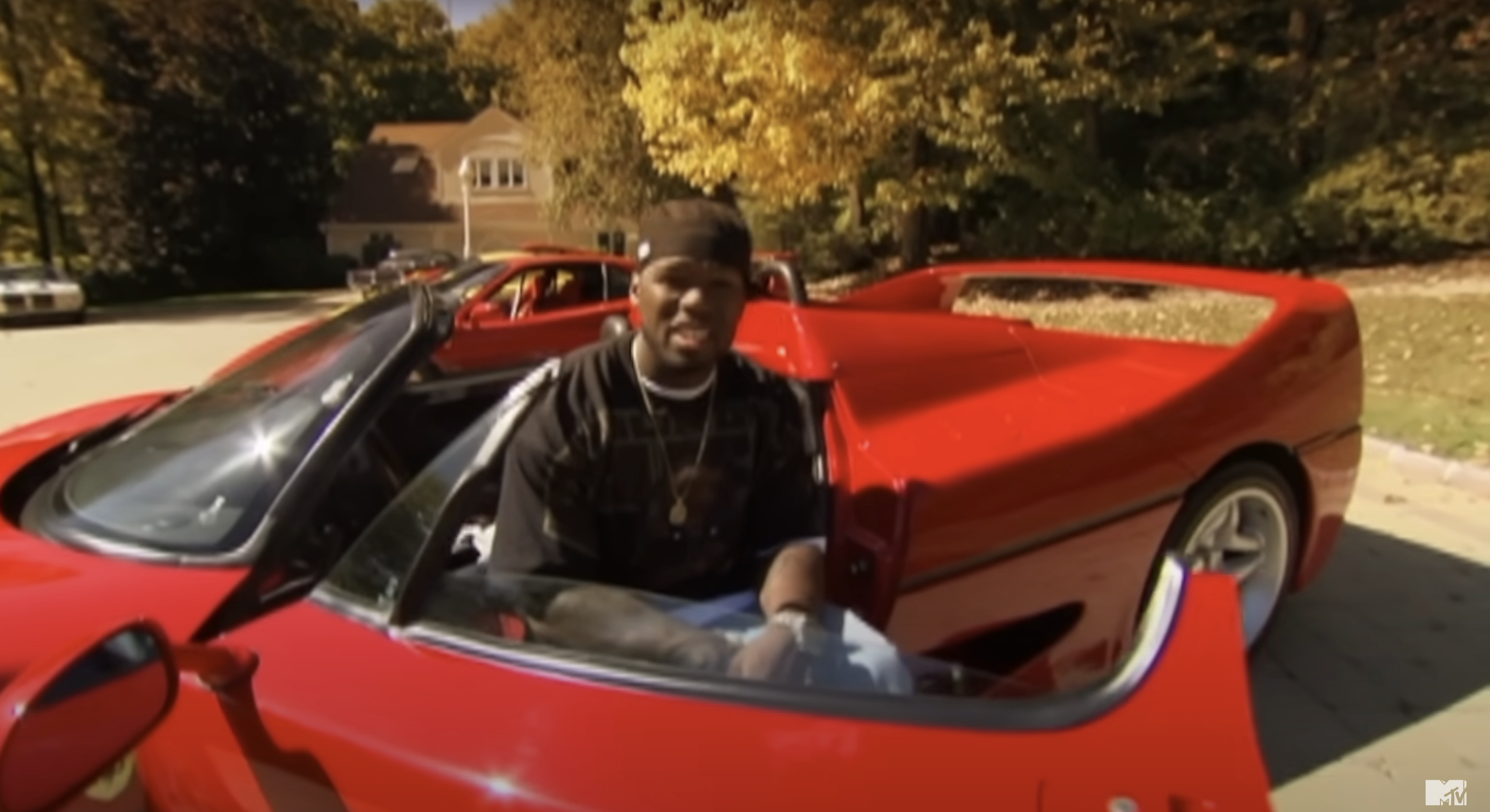 50 Cent sitting in a sports car, smiling, with another similar car next to him