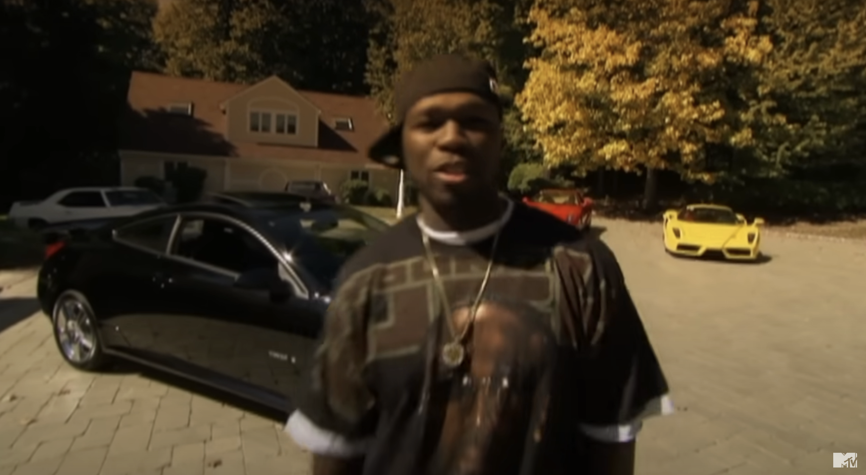 50 Cent standing by a car, wearing a cap and graphic tee, with two sports cars in the background
