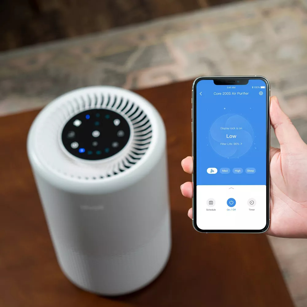 Hand holding smartphone with air purifier app open, air purifier device in the background on floor