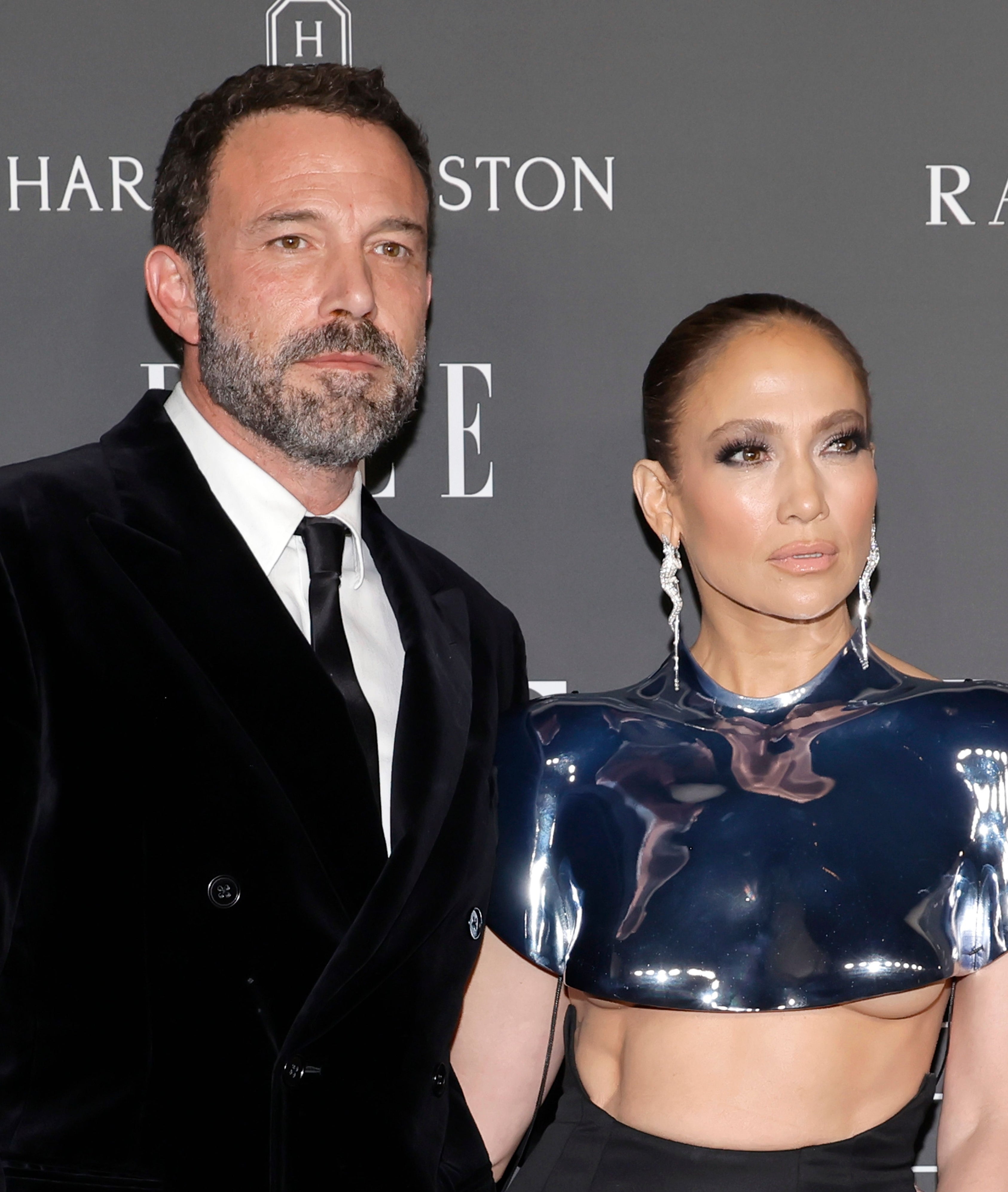 Ben Affleck and Jennifer Lopez posing together; he in a black suit, she in a metallic crop top