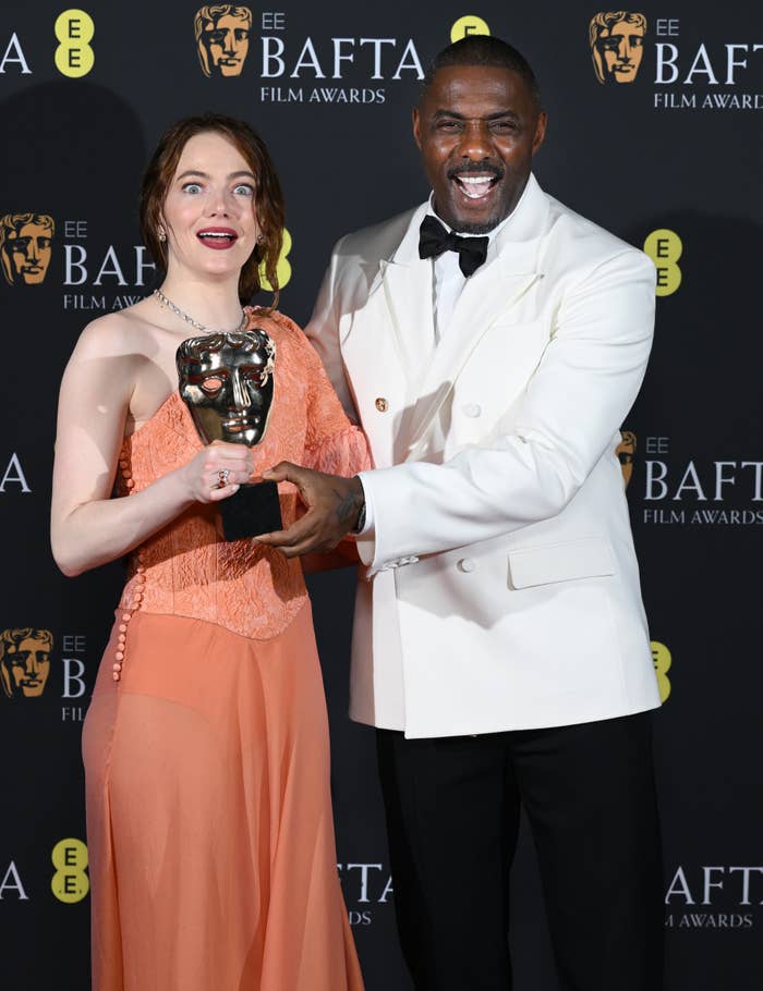 Emma in an orange gown holding a BAFTA award with Idris Elba, in a white tuxedo