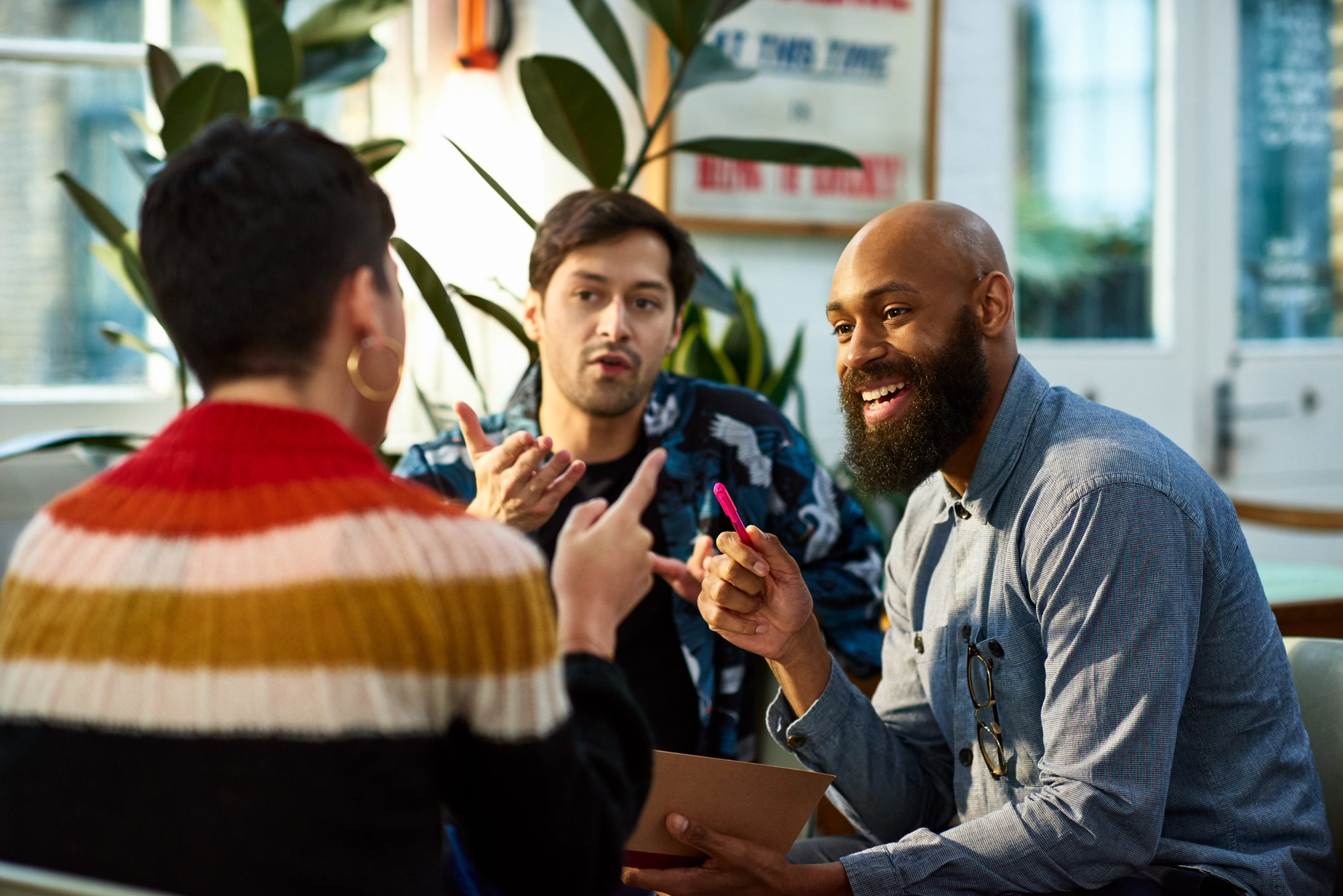 Three people in a casual meeting, two listening to one who is gesturing while speaking
