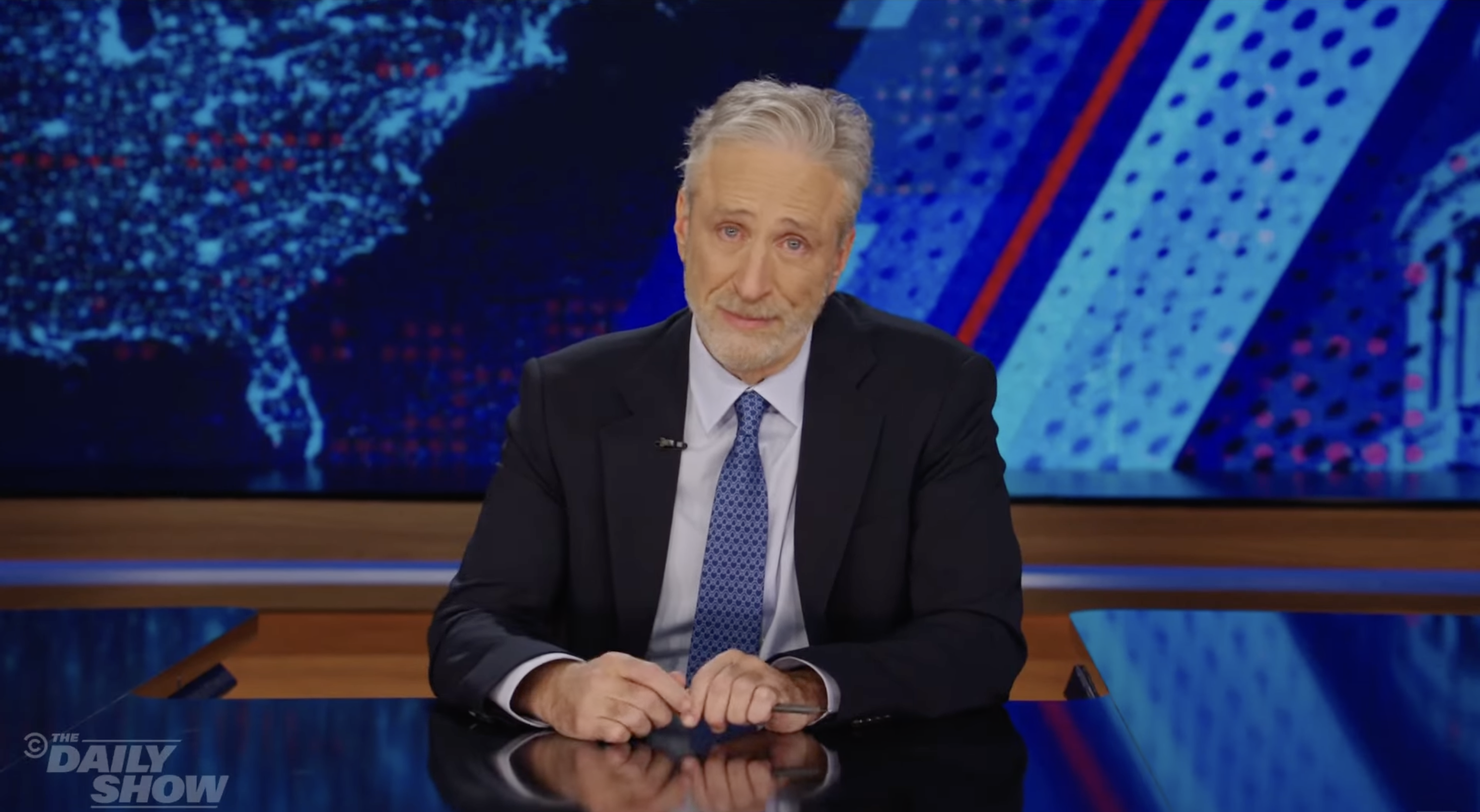 Jon Stewart in a suit hosts &quot;The Daily Show,&quot; sitting at a desk with the show&#x27;s graphics in the background