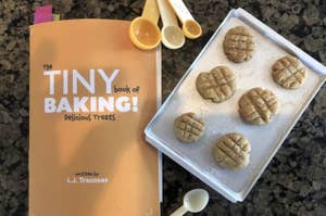 a rveiewer photo of a. tiny cooking sheet topped with tiny cookies next to a book titled "The tiny book of baking"