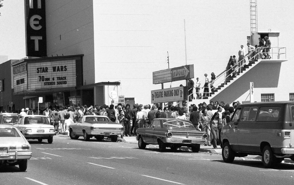 Crowd lining up outside a theater for &quot;Star Wars&quot; premiere, classic cars parked in front