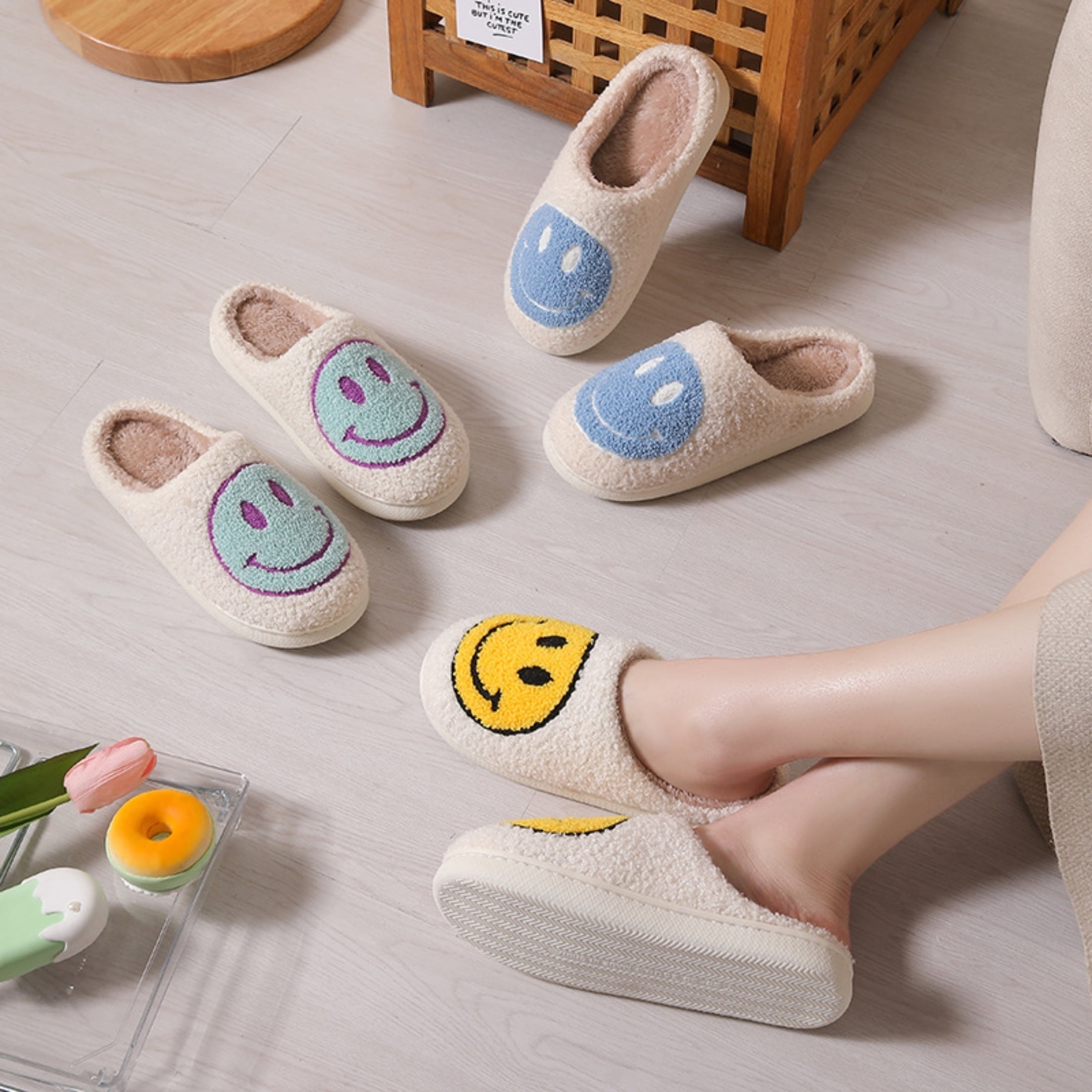 A person&#x27;s feet wearing one smiley face slipper, with other slippers nearby on a floor