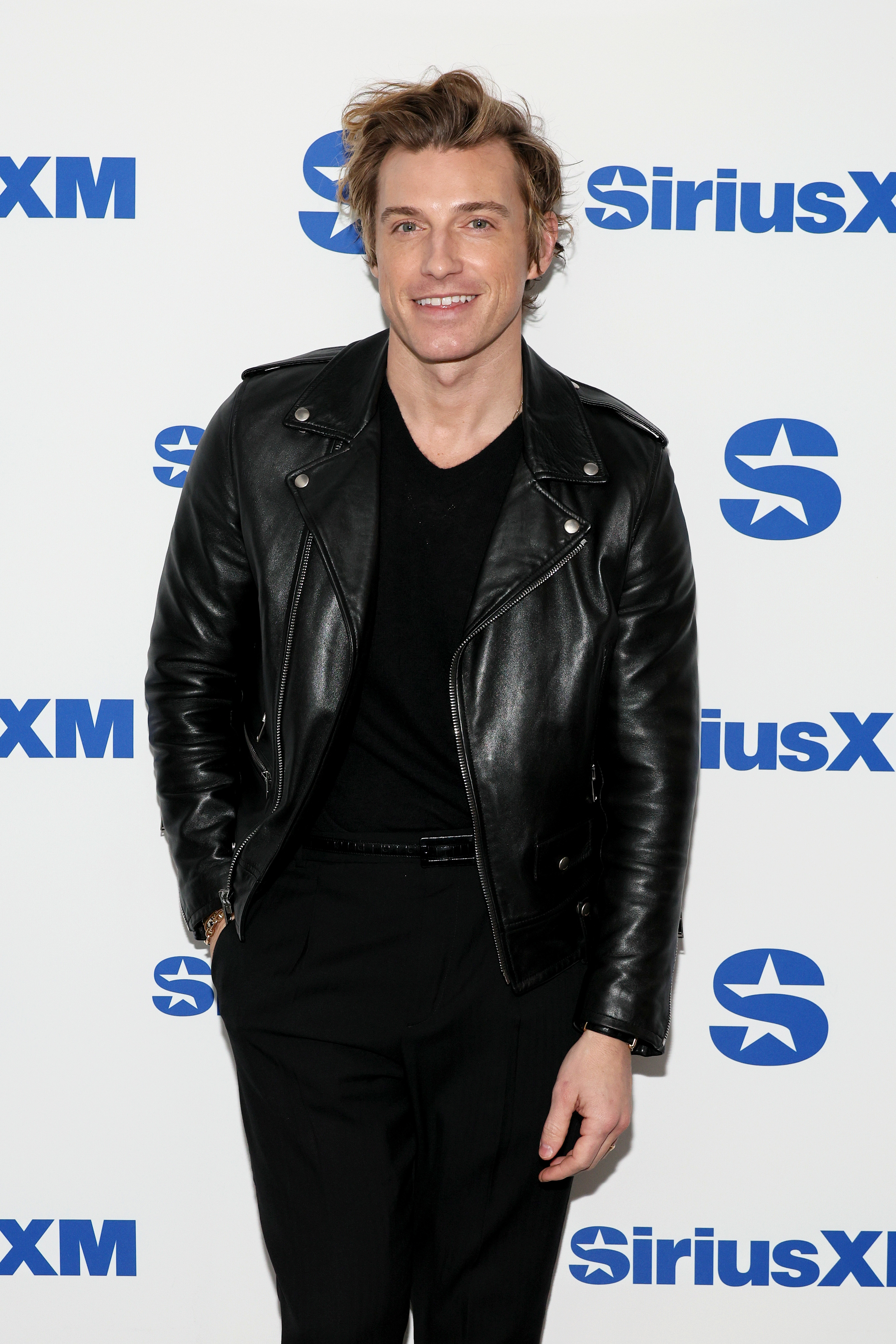 Jeremiah in a leather jacket and attire smiles standing before a SiriusXM backdrop
