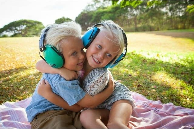 Two children wearing noise cancelling headphones embrace on a picnic blanket outdoors