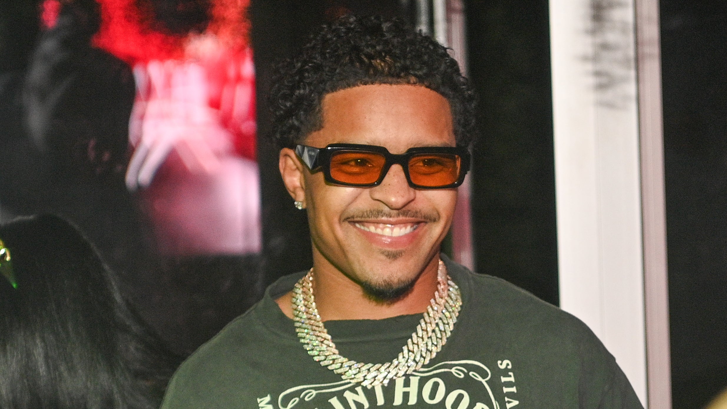 Artist smiling in a graphic tee, layered necklaces, and tinted sunglasses at a music event