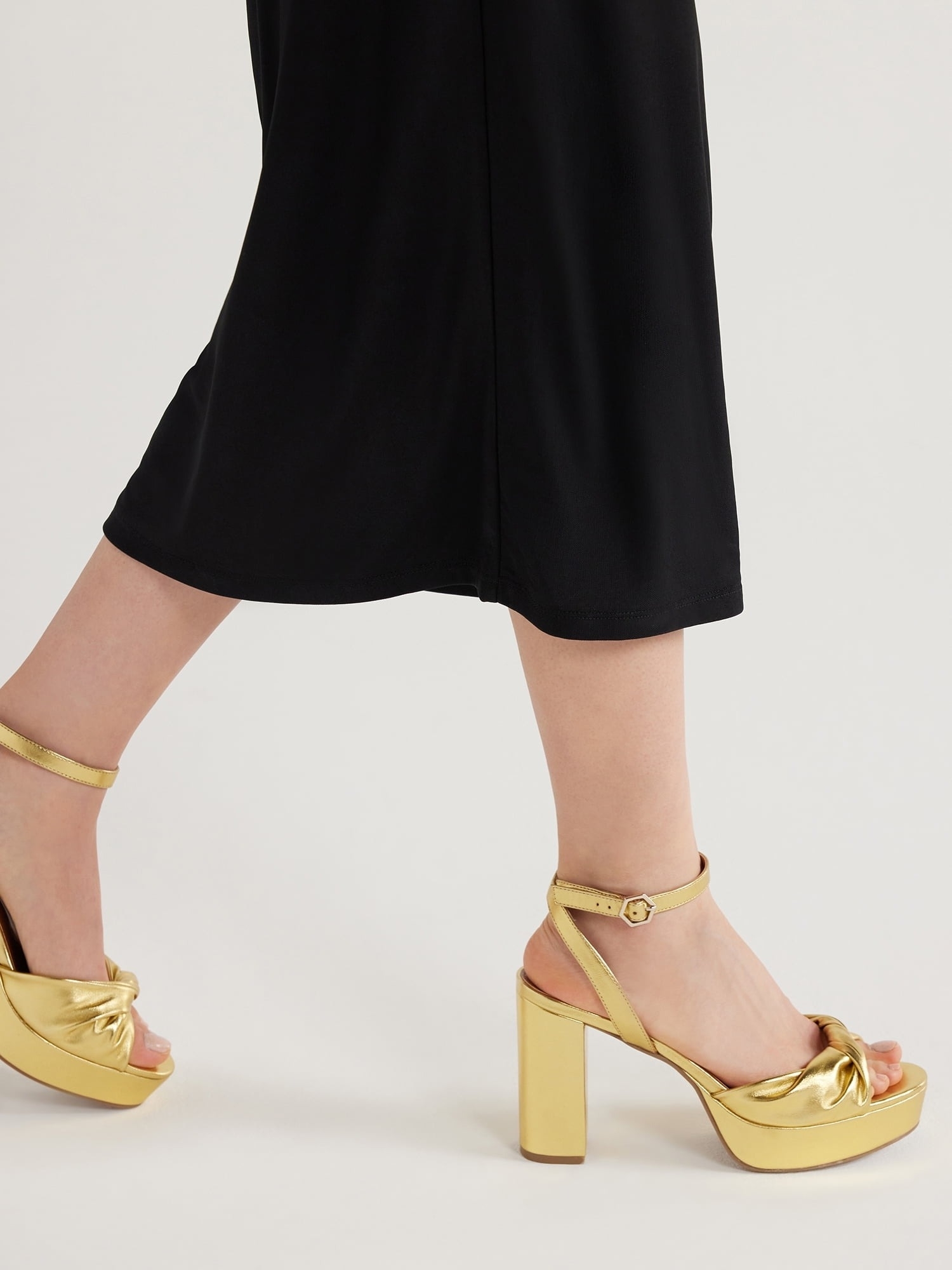 Close up of a model wearing gold high-heeled sandals with a bow detail, paired with a black dress