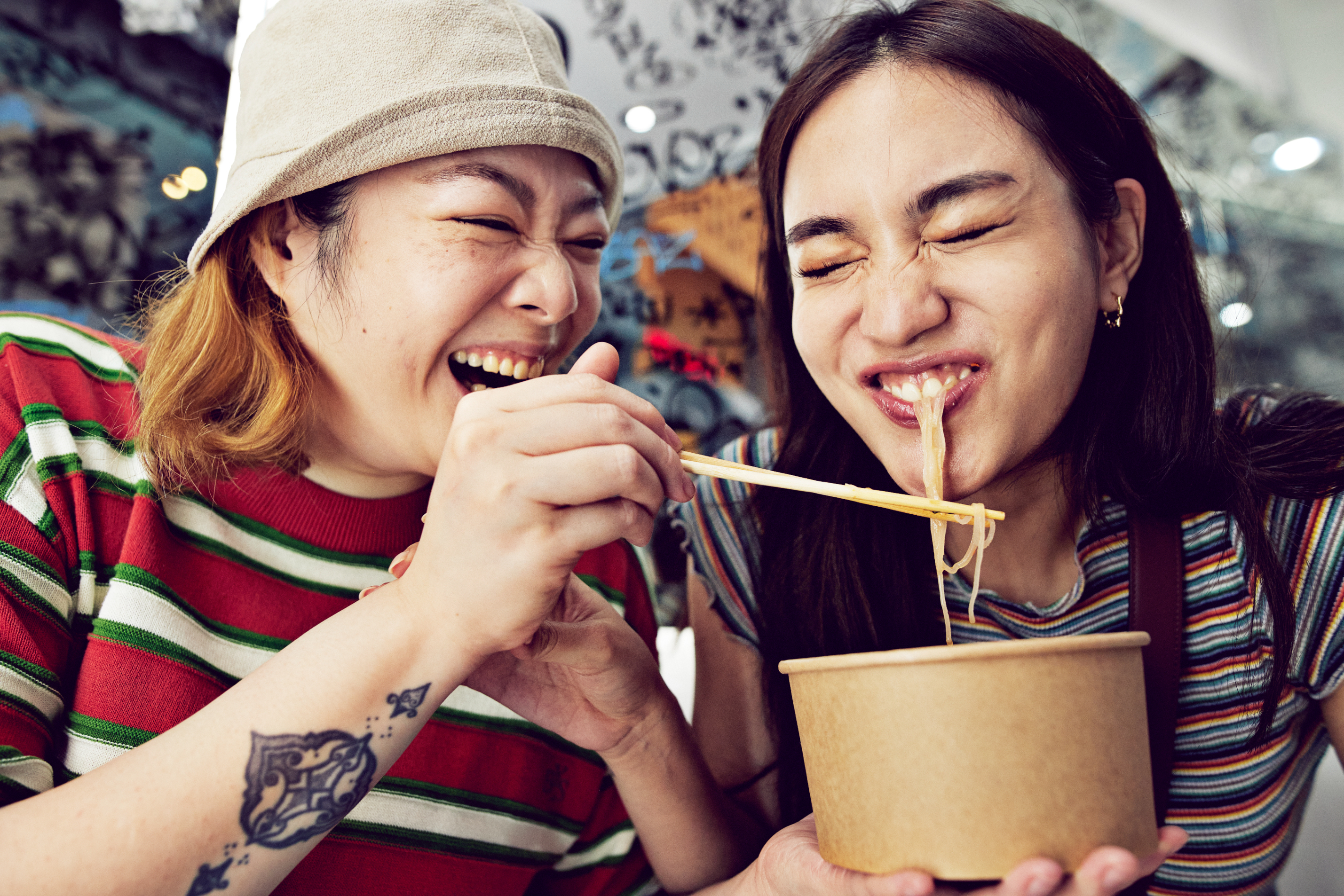 Two people are sharing a laugh while eating noodles from a takeout container