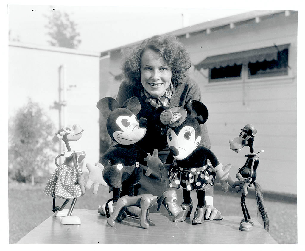 Woman with curly hair poses behind table with Mickey and Minnie Mouse figures, plus other Disney character toys