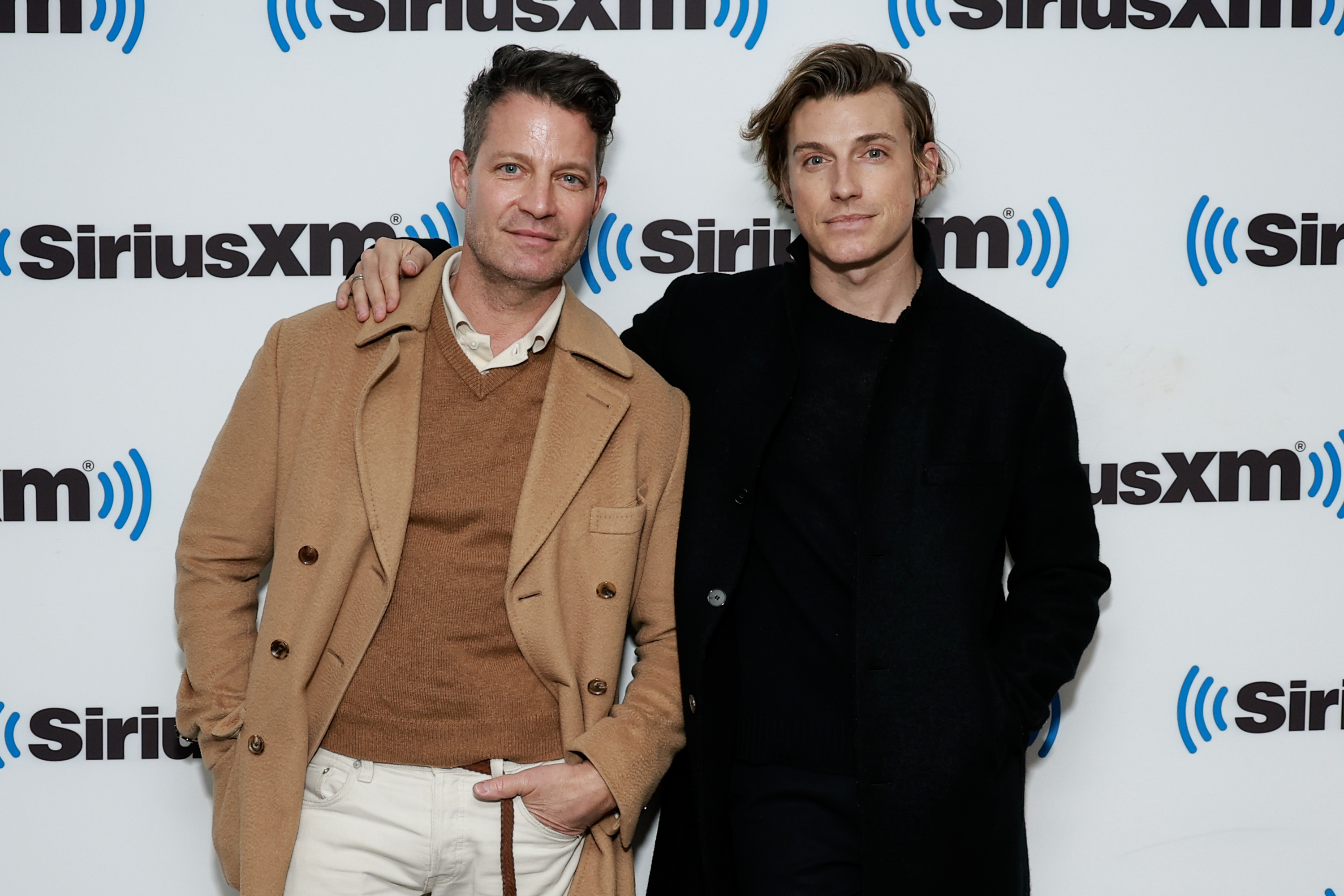 Two men posing at a SiriusXM event, one in a tan coat and white shirt, the other in a black outfit