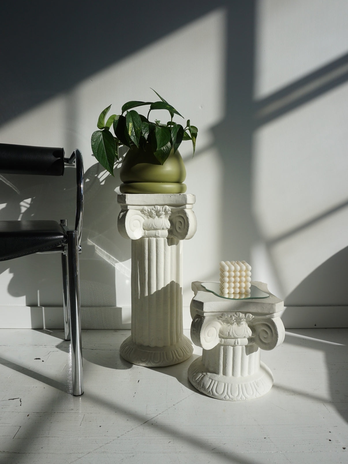 Two classical-style pedestals with a potted plant and decorative beads in a shadow-patterned room