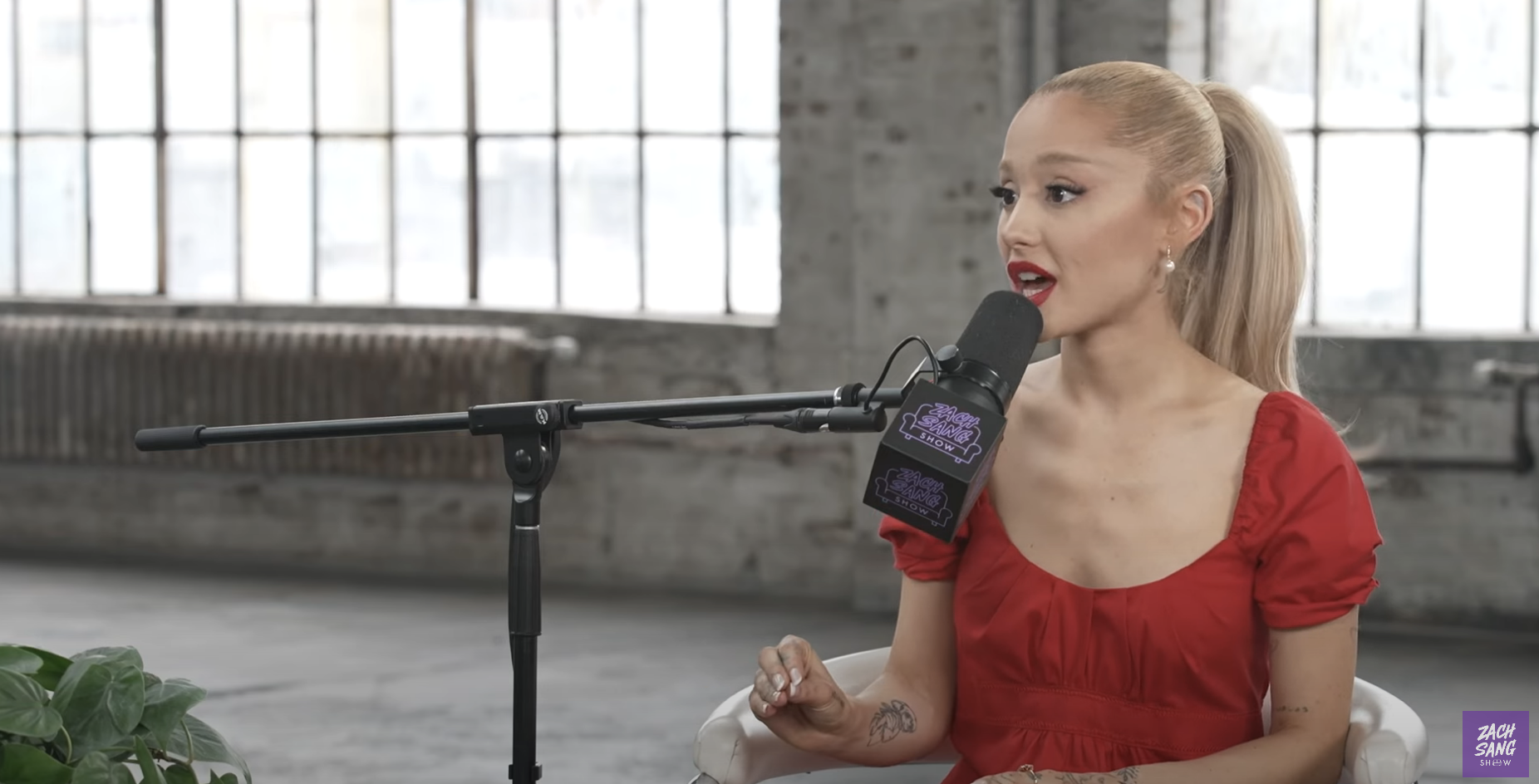 Ariana Grande in a red dress speaking into a microphone during an interview