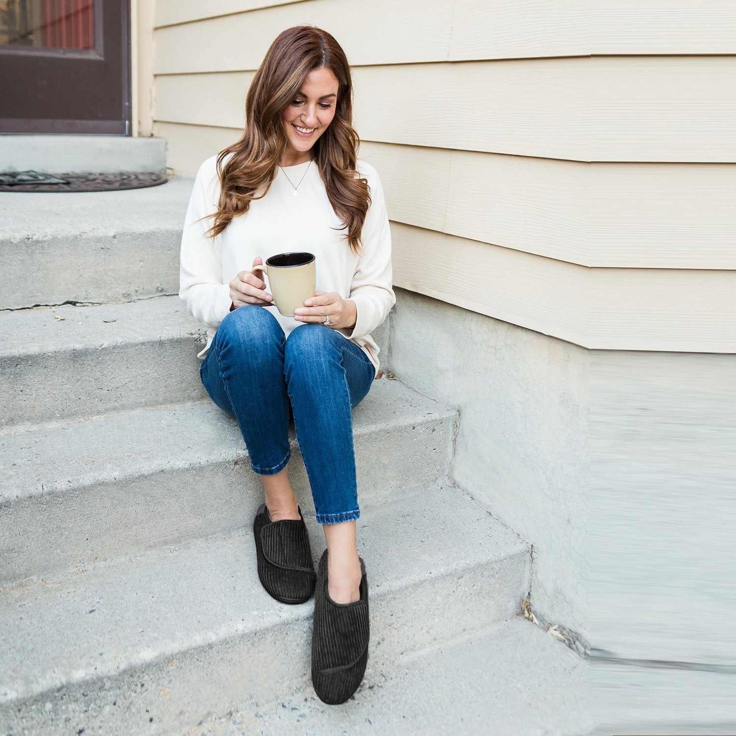 model sitting on steps holding a mug, wearing the black slippers, a white top and blue jeans