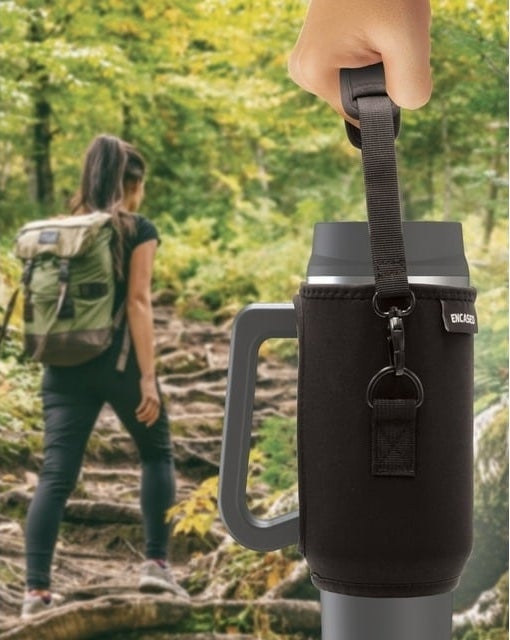 Person hiking in the background, another&#x27;s hand holding a travel mug in the foreground