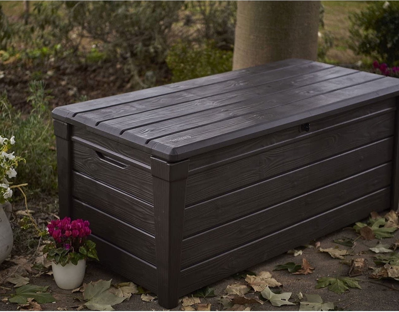 dark brown outdoor storage chest with a closed lid, placed on a patio next to a potted plant