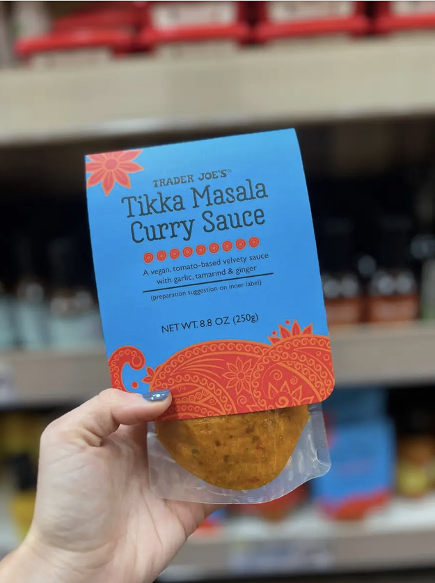Hand holding a jar of Trader Joe&#x27;s Tikka Masala Curry Sauce in front of grocery shelves