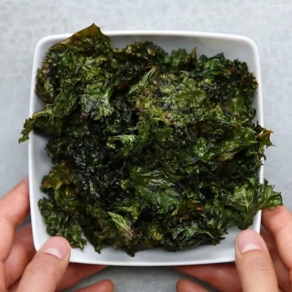Hands holding a square bowl filled with crispy kale chips