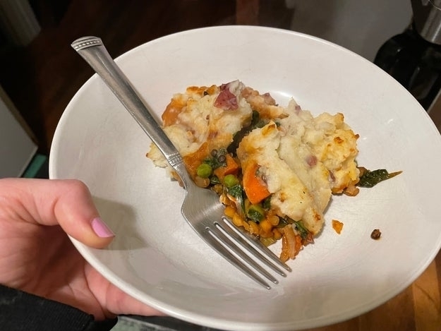 A person&#x27;s hand holding a plate with a portion of shepherd&#x27;s pie, including vegetables and mashed potatoes