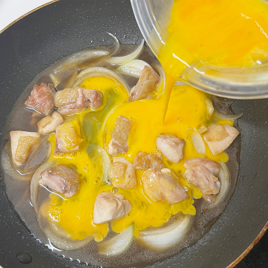 フライパンで玉ねぎと一緒に調理されている鶏肉に卵液を注ぐ様子。