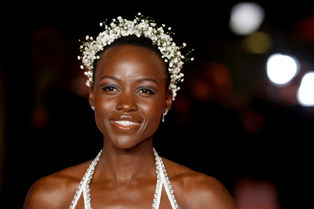 Lupita Nyong&#x27;o smiling in a jeweled dress with flower headpiece at a celebrity event