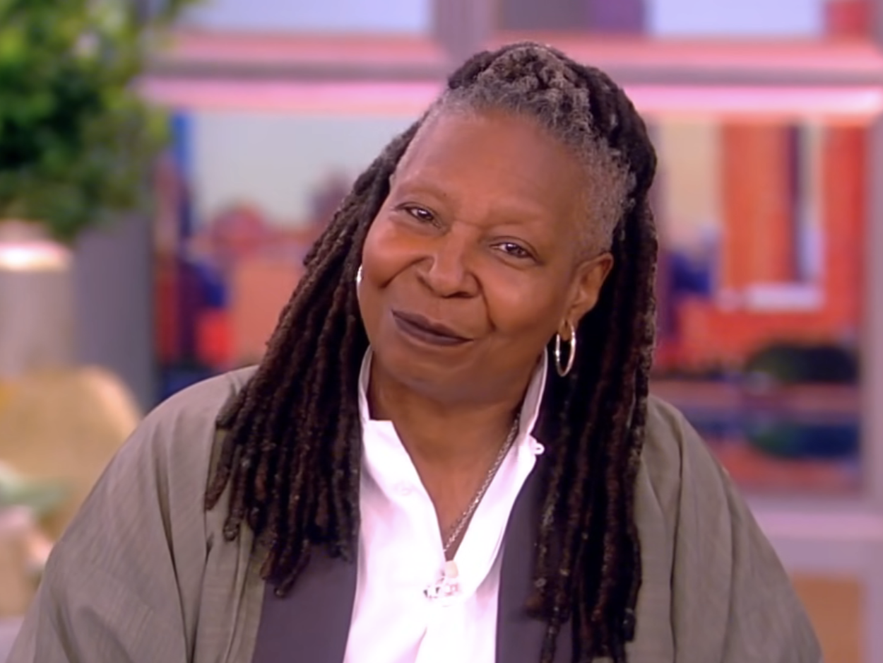 Whoopi Goldberg smiles in a studio, with her hair in long locs