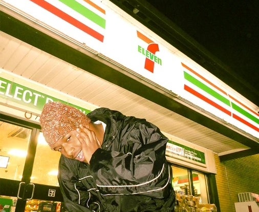 Person posing with hand gesture in front of 7-Eleven, wearing a shiny jacket, at nighttime