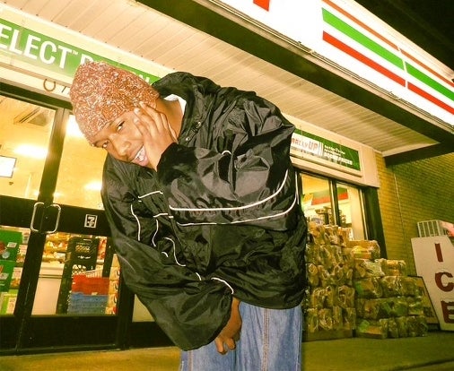 Person posing with hand gesture in front of 7-Eleven, wearing a shiny jacket, at nighttime