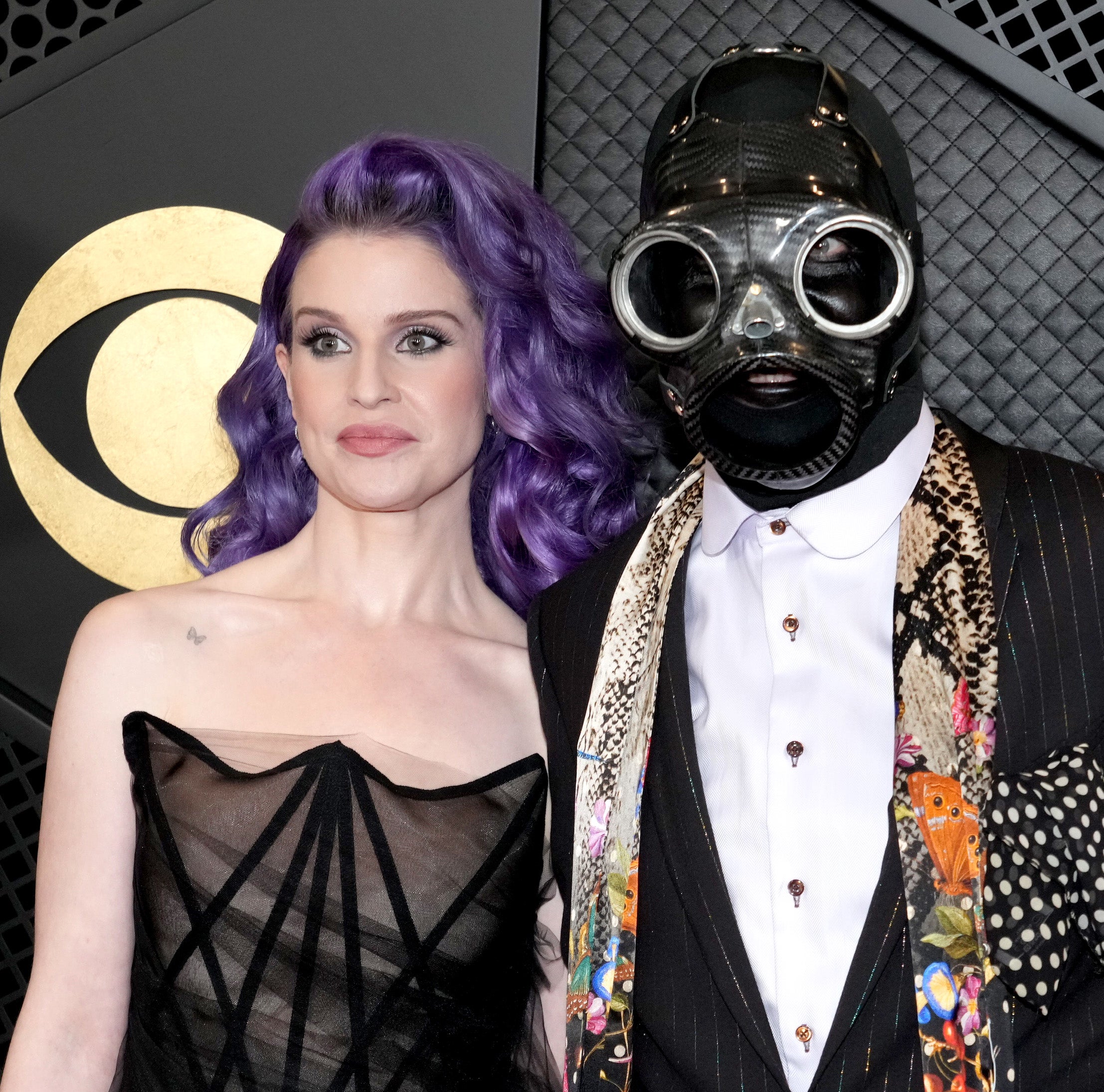 Two individuals posing, one in a black dress and purple hair, the other in a suit with a patterned design and wearing a gas mask
