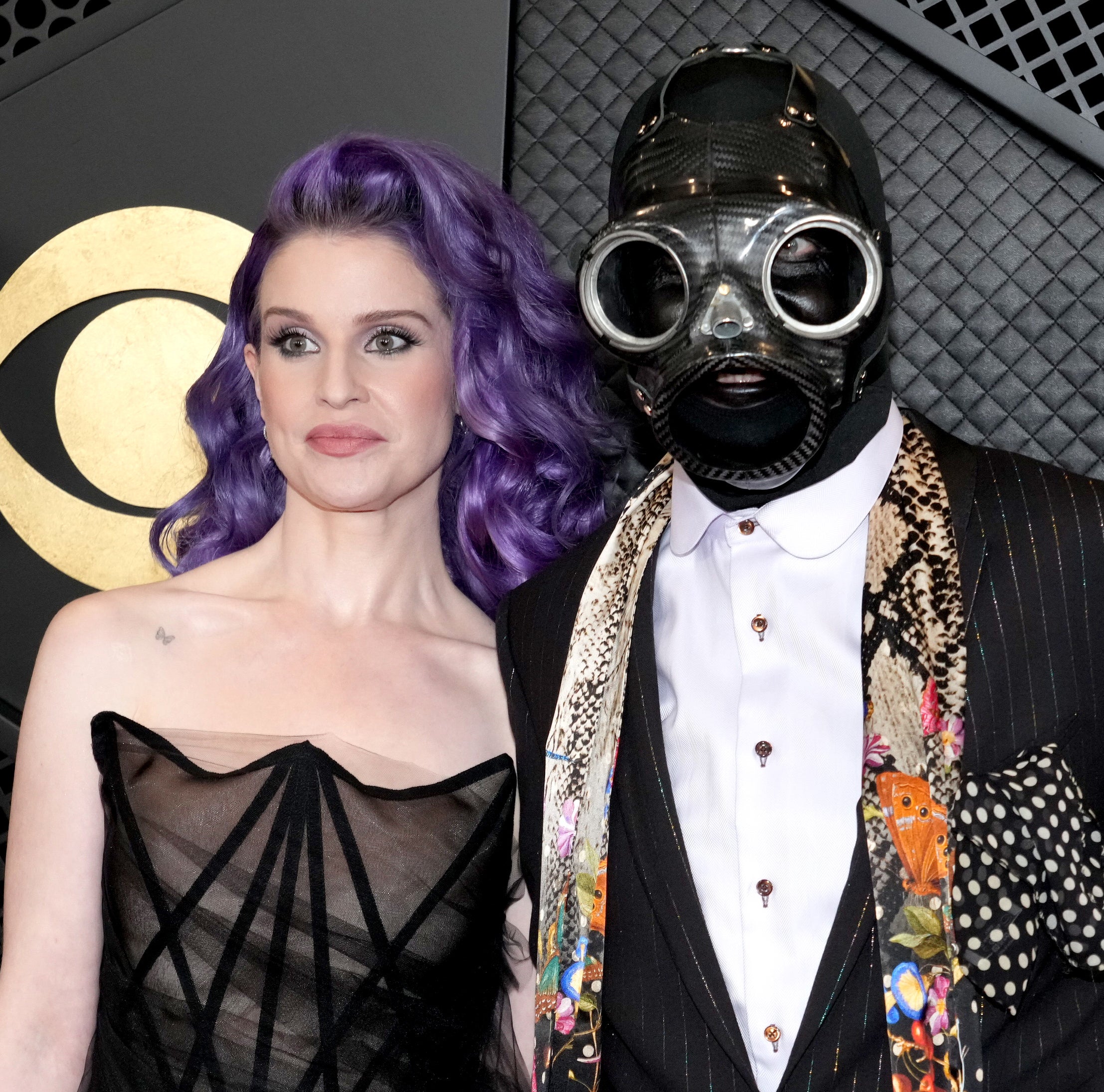 Two individuals posing, one in a black dress and purple hair, the other in a suit with a patterned design and wearing a gas mask