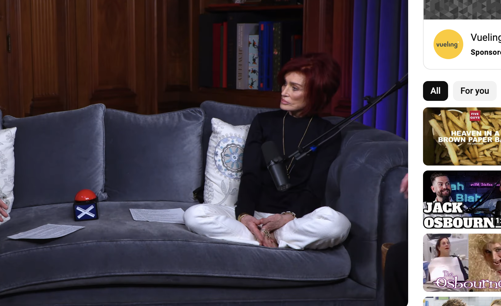 Two people seated in an interior set for an interview, with one wearing a dark outfit and the other in a red and black ensemble