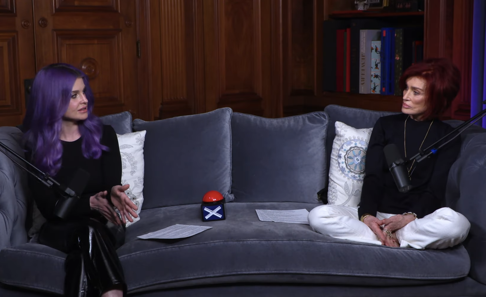 Two people seated in an interior set for an interview, with one wearing a dark outfit and the other in a red and black ensemble