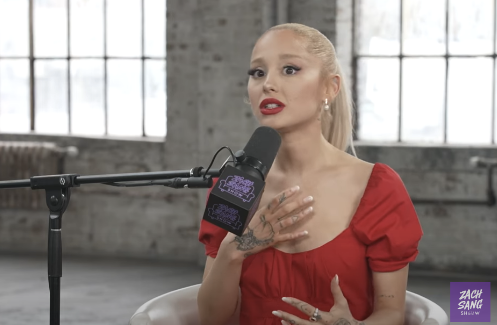 Ariana Grande in a puff-sleeve dress gestures while speaking into microphone during an interview