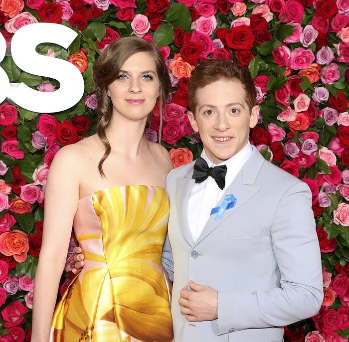 Lilly Jay and Ethan Slater posing in front of a flower wall; one in a floral gown, the other in a light suit