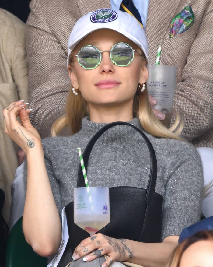 Ariana Grande in visor and sunglasses holding a drink at an event. She wears a sweater top and accessories