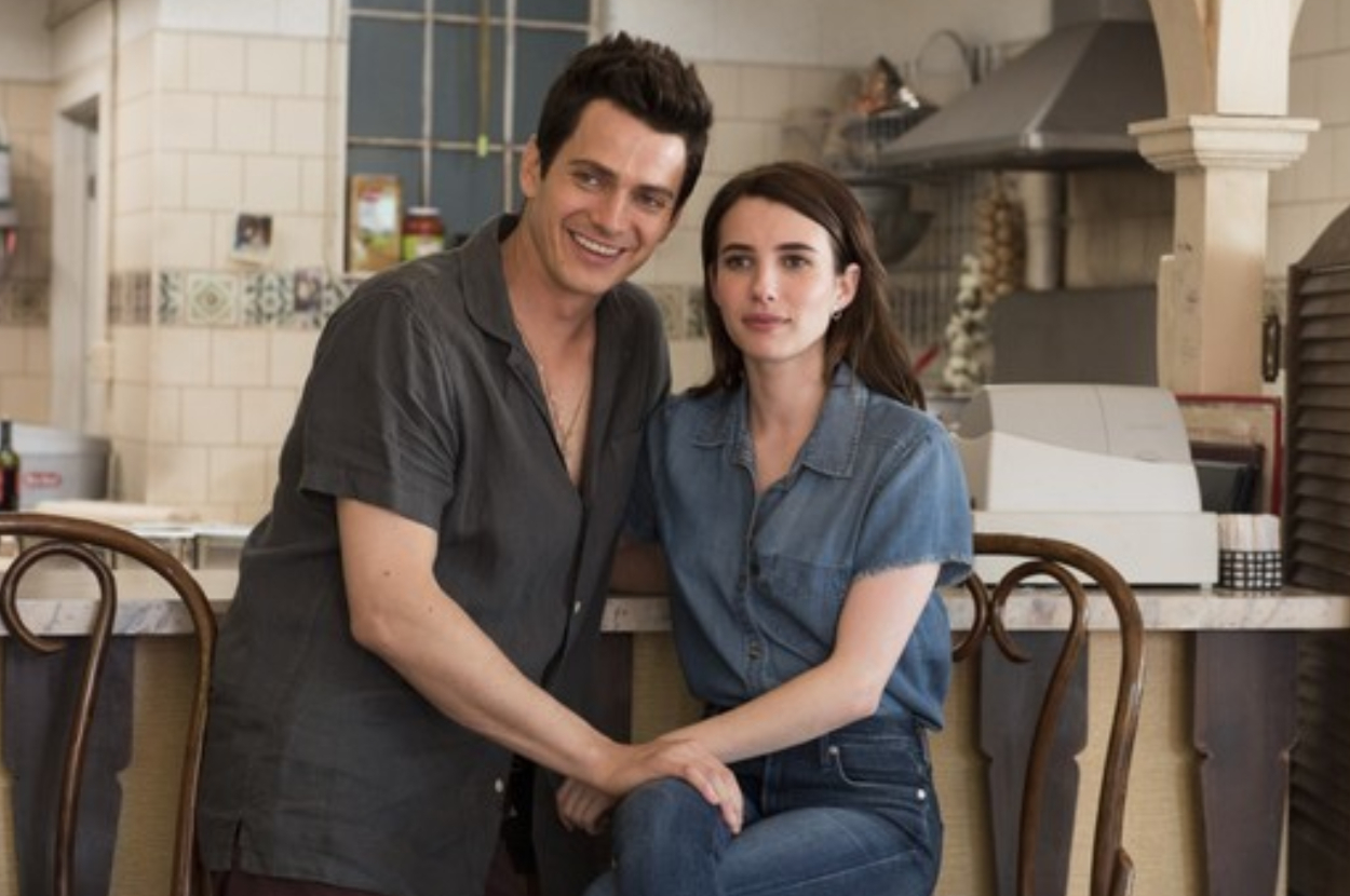 Two actors portraying a couple, seated in a diner, smiling at the camera. They wear casual denim outfits