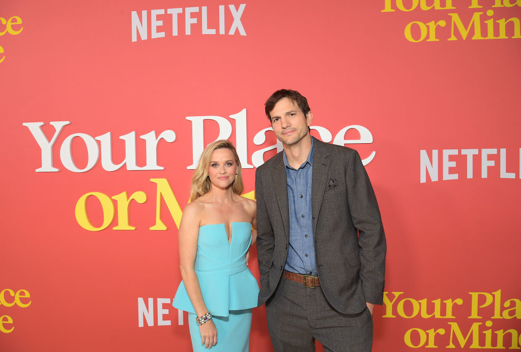 Two people standing in front of a promotional backdrop. The person on the left is wearing a strapless blue outfit, and the person on the right is in a gray suit