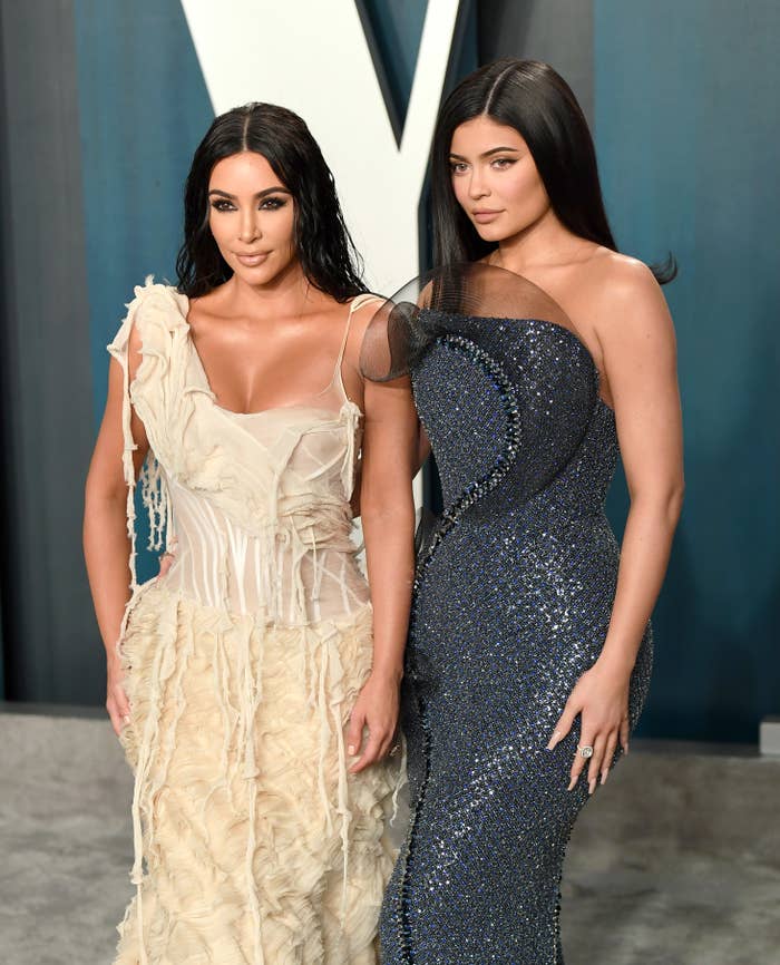 Kim Kardashian and Kylie Jenner posing in elegant gowns at an event. Kim is in a ruffled dress; Kylie in a sequined gown