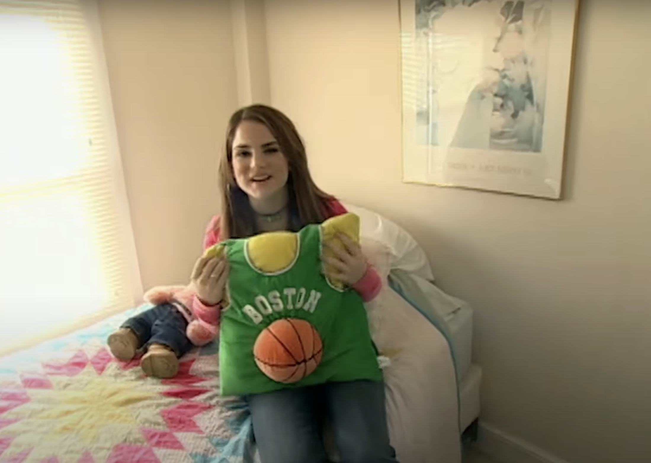 JoJo sitting on bed holding a sports-themed pillow, with stuffed toys behind her