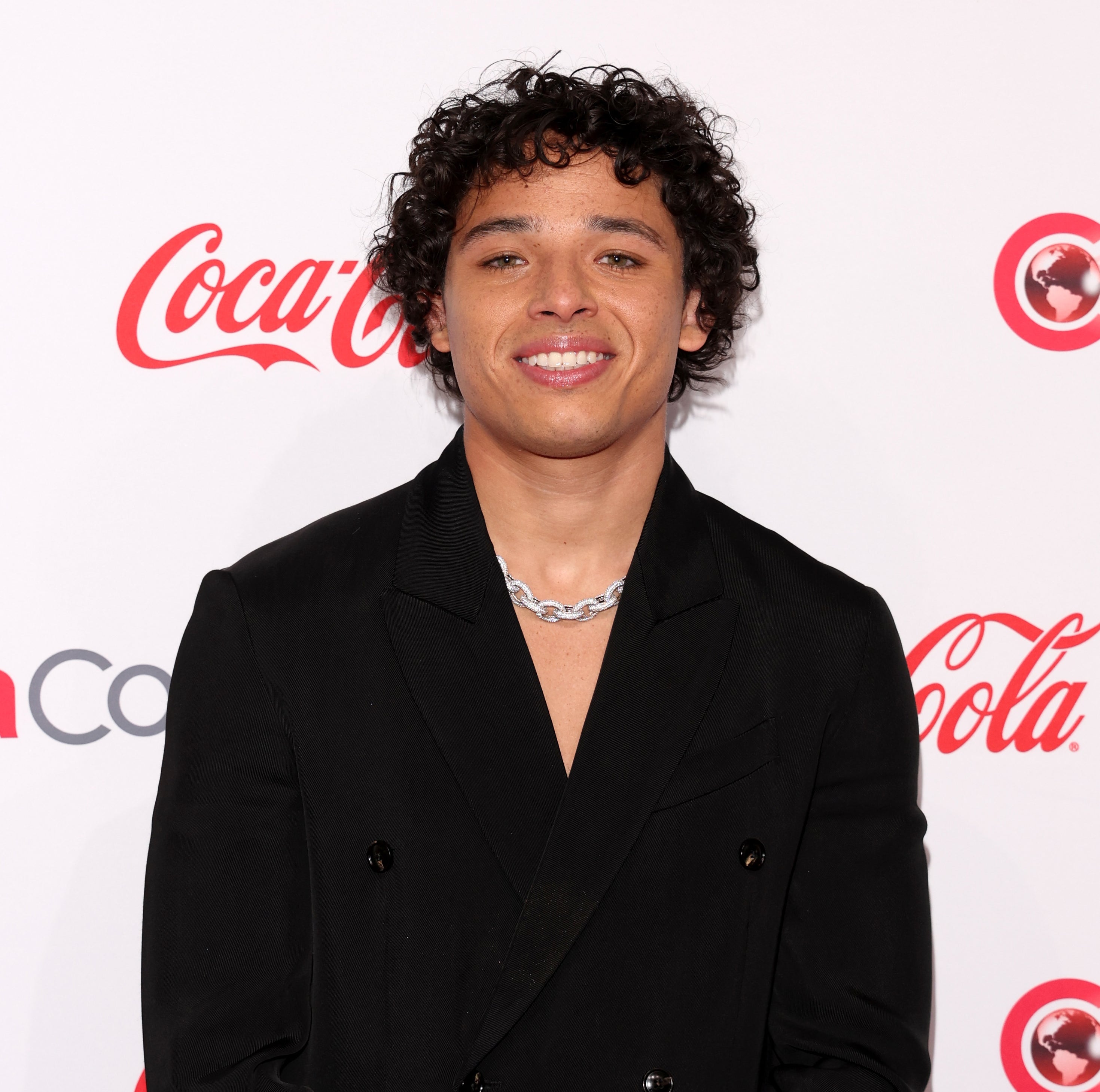 Anthony Ramos in a black jacket and jeweled necklace smiling on the CinemaCon red carpet