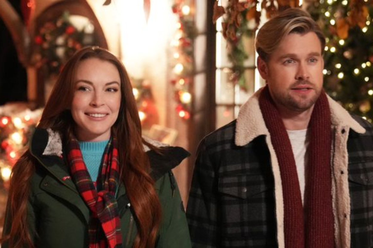 Two actors from a holiday-themed movie, dressed in warm winter clothes, standing with festive decorations in the background