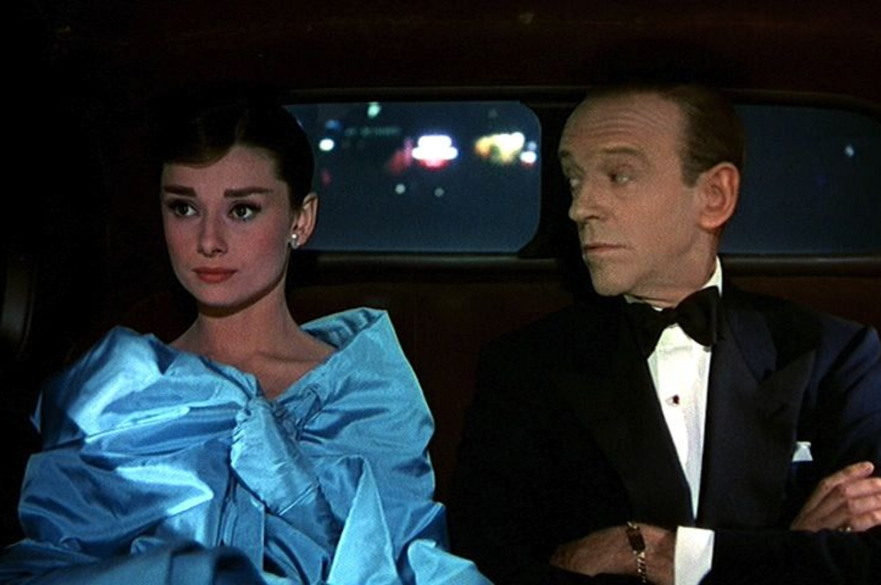 Audrey Hepburn and actor in formal attire sitting in a car from a classic movie scene