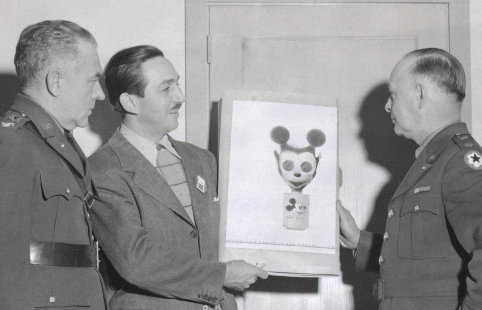 Three men in military uniforms looking at a Mickey Mouse gas mask sketch