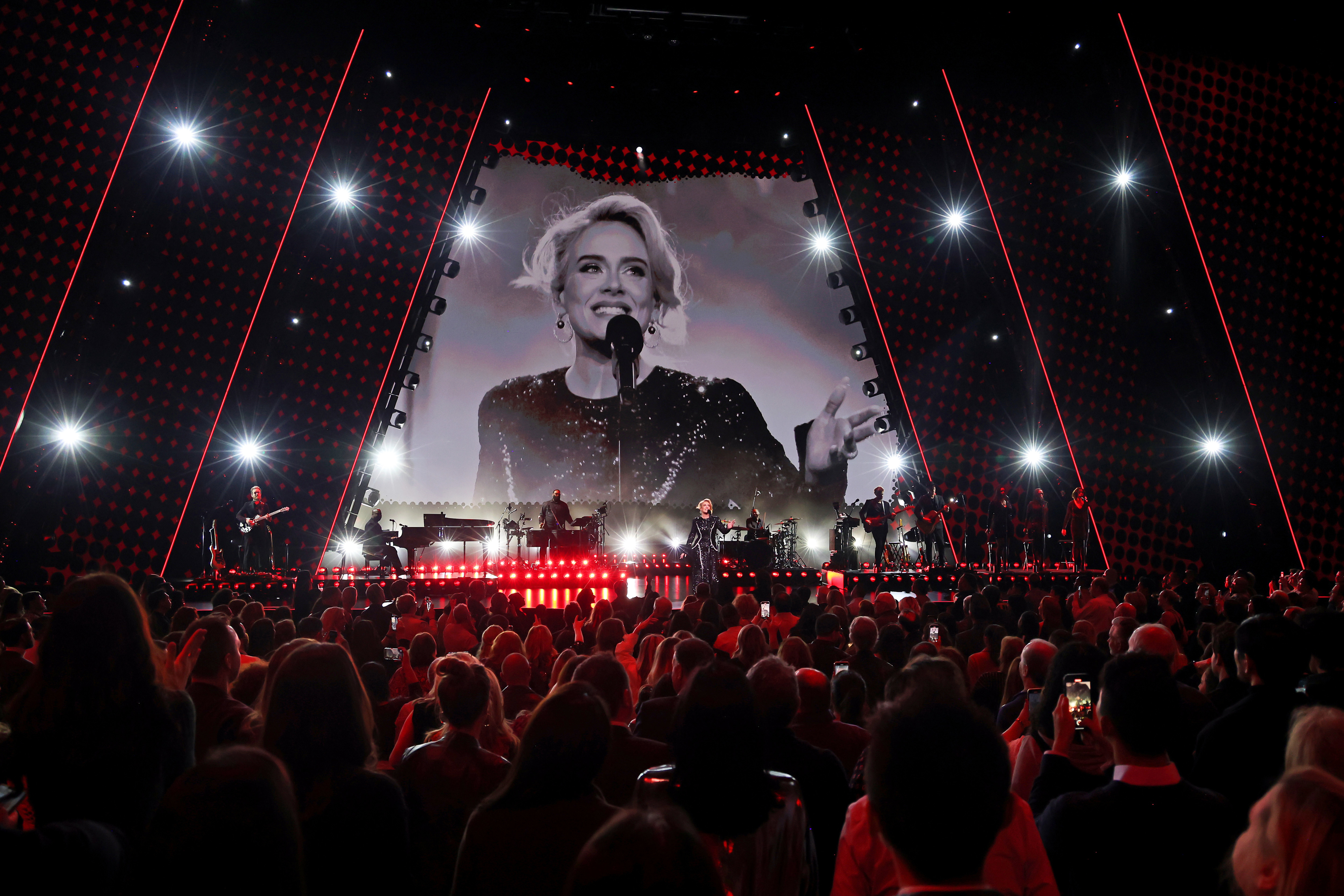 Crowd watching Adele perform on stage with a band, large screen in the background showing the artist